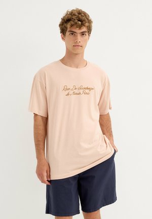 Joven con cabello rizado que lleva una camiseta rosa claro con texto marrón en cursiva y pantalones cortos azul marino oscuro, de pie contra un fondo liso.