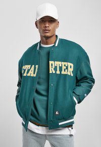 Teal varsityjack met gele letters, witte accenten en een zachte textuur. Gemaakt over een bijpassende sweatshirt, aangevuld met lichte jeans.