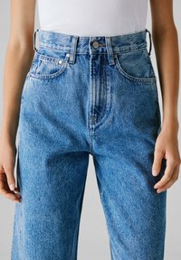 Persona vistiendo jeans de mezclilla de tiro alto, de color azul claro, y una blusa sin mangas blanca metida por dentro, de pie con las manos relajadas a los lados.