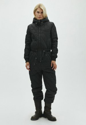 Femme aux cheveux blonds courts portant une combinaison matelassée noire à capuche et des bottes noires, debout devant un fond blanc uni.
