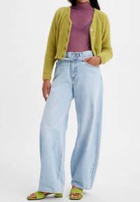 Grön fluffig kofta, lila mock neck-top, ljus denim byxor med vida ben och gröna klacksandaler. Påtagliga texturer och livliga färger.