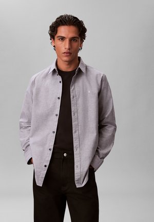 Jonge man met krullend haar draagt een lichtgrijs overhemd met knopen over een zwart shirt, handen in de zakken, staand tegen een effen grijze achtergrond.