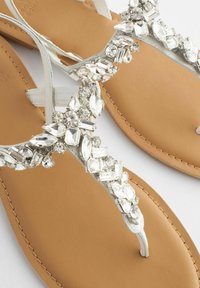 Kristalversierde teensandalen met een witte band, voorzien van een beige zool en verschillende heldere kristalvormen als decoratie.