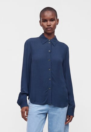Frau mit kurzen Haaren, die eine marineblaue Bluse mit Knöpfen und hellblaue Jeans trägt, steht vor einem schlichten Hintergrund.