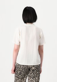 Osoba s krátkými černými vlasy, oblečená v bílém vzorovaném tričku s krátkým rukávem a kalhotách se zebřími pruhy v černobéžové barvě, otočená zády.