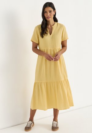Kvinde iført en tekstureret gul midi-kjole med korte ærmer og lagdelt nederdel samt brune sneakers, stående foran en hvid baggrund.