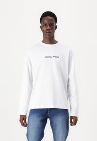 Langermet hvit bomull T-skjorte med "RED BULL RACING" i svart tekst, kombinert med blå denimjeans. Minimalistisk design, standard passform.