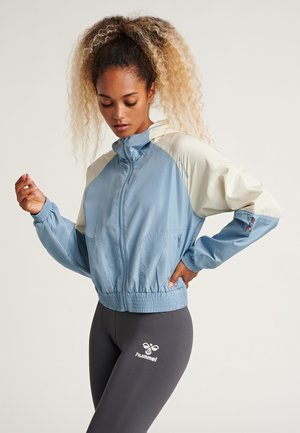 Veste coupe-vent bleu clair avec des accents crème, des poignets et un ourlet élastiques, des poches avant et une fermeture éclair. Associée à un legging gris ajusté.