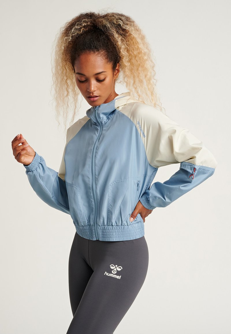 Light blue windbreaker jacket with cream accents, elastic cuffs and hem, front pockets, and a zip-up closure. Paired with fitted gray leggings.