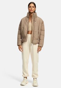 Opgeblazen beige jas met hoge kraag, ritssluiting aan de voorkant en zijzakken; gedragen over een lichtbeige crop top en crème sweatpants met een losse pasvorm.
