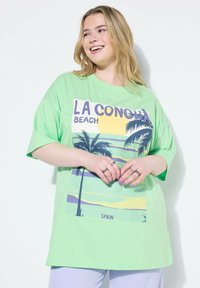 Mujer sonriendo con cabello rubio, vistiendo una camiseta verde suelta con un estampado tropical de "La Concha Beach España" y pantalones de color claro.