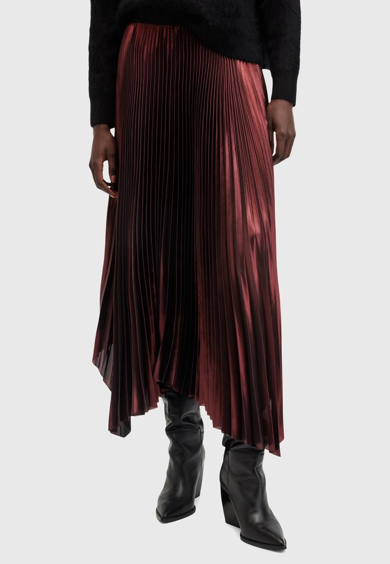 Personne portant une jupe asymétrique plissée brillante couleur bordeaux, des bottes à talons noires et un pull noir duveteux, debout sur un fond uni.