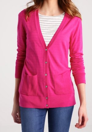 Donna che indossa un cardigan rosa acceso abbottonato sopra una maglia a righe grigia e bianca con jeans blu, in piedi con le braccia rilassate.