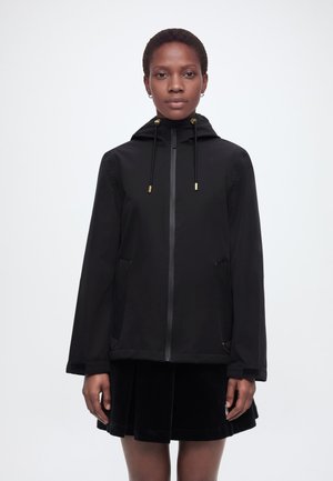 Kobieta z krótkimi włosami, ubrana w czarną bluzę z kapturem na zamek i czarną plisowaną spódnicę, stojąca przodem na jednolitym tle.