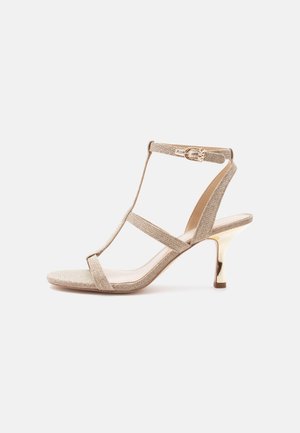 Beige stroppesandal til kvinder med guldfarvet lav hæl og ankelspænde, vist på hvid baggrund.