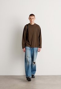 Chemise marron à manches longues, jean bleu clair déchiré avec des rips, et chaussures noires. Le modèle se tient devant un fond neutre, centré dans l'image.