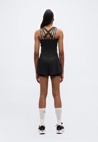 Γυναίκα που στέκεται με πλάτη, φορώντας μαύρο αθλητικό τοπ με διασταυρωμένα λουριά, μαύρα στρωμένα σορτς, λευκές κάλτσες Puma και μαύρα αθλητικά παπούτσια σε απλό φόντο.