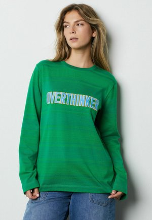 Green long-sleeve t-shirt featuring "OVERTHINKER" in blue lettering. Smooth texture with a crew neck and relaxed fit, paired with blue jeans.