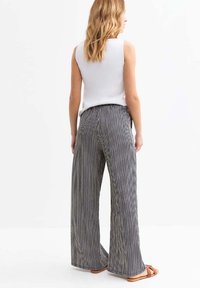 Una donna sta in piedi con le spalle alla telecamera, indossando un top bianco senza maniche e pantaloni a gamba larga a righe bianche e nere.