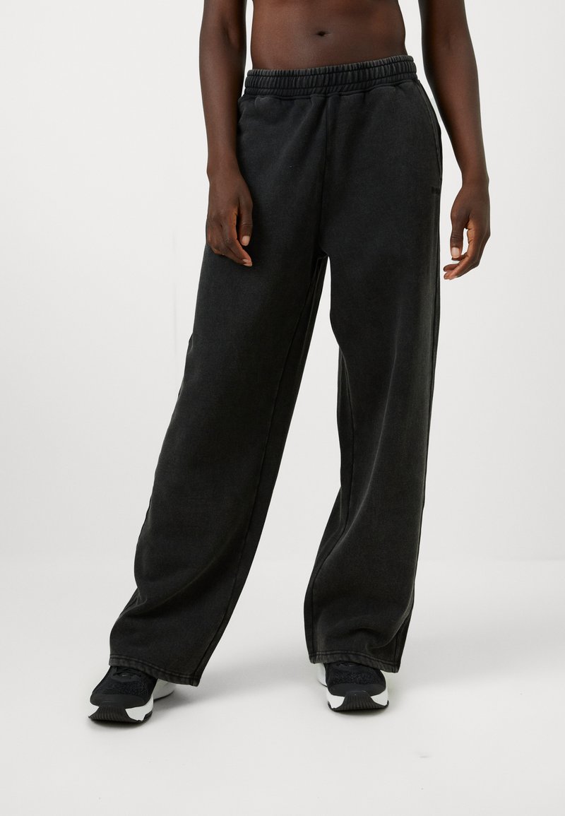 Juodos plataus kirpimo sportinės kelnės su elastinga juosme, pagamintos iš minkštos medžiagos, pasižyminčios lygiu paviršiumi ir minimaliais siuvimo elementais.