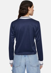 Maglietta a maniche lunghe di colore blu navy con strisce verticali, texture a costine e colletto bianco con strisce blu navy. Indossata con pantaloni cargo blu chiaro.