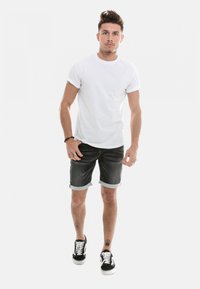 T-shirt en coton blanc, avec des manches retroussées, associé à un short en denim foncé avec des ourlets. Baskets noires avec des lacets blancs et un tatouage visible sur le bras.