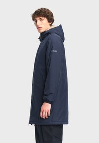 Marineblauwe gewatteerde parka met ritsluiting, elastische manchetten en een fleece-gevoerde binnenkant. Heeft een logodetail op de mouw en een rechte snit.
