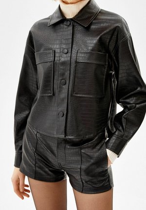 Frau trägt eine schwarze pseudo-lederne strukturierte Hemdjacke mit großen Brusttaschen und passende hoch taillierte Shorts über einem cremefarbenen Rollkragenpullover.