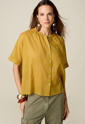 Femme portant un chemisier à manches courtes moutarde boutonné, un pantalon vert olive avec des poches, de grandes boucles d'oreilles et des bracelets épais au poignet gauche.