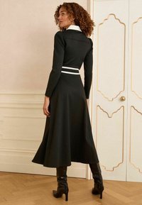 Vestido negro con un cuello y cinturón blancos, mangas largas, falda acampanada y tejido texturizado. Llevado con botas negras hasta la rodilla.