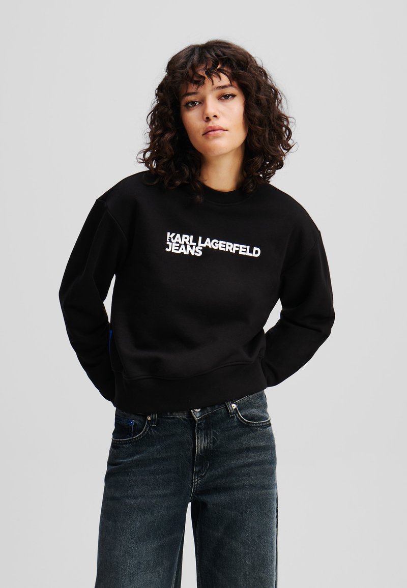 Black sweatshirt with white "KARL LAGERFELD JEANS" text, crew neck, dropped shoulders, and ribbed cuffs, paired with dark denim jeans.