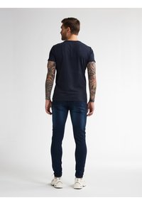 Námornícky modré tričko s krátkým rukávem, úzký střih, nošené s tmavými džíny. Model má tetování a stojí zády k divákovi.