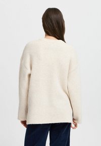 Pull en tricot côtelé beige surdimensionné avec un col rond, des manches longues et un ourlet fendu. Texture douce, variation de couleur subtile visible.