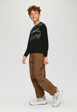 Ein rothaariger Junge trägt einen schwarzen Grafikpullover, braune Hosen und weiße Turnschuhe, steht vor einem schlichten hellen Hintergrund und lächelt leicht.