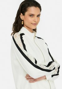 Veste blanche avec des rayures noires et des accents en dentelle sur les manches, dotée d'une fermeture éclair dorée et d'un col haut. Texture lisse.