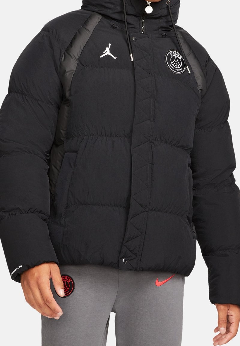 Doudoune noire avec les logos Paris Saint-Germain et Jumpman, portée avec un pantalon gris avec un patch rouge Paris Saint-Germain et un swoosh rouge Nike.