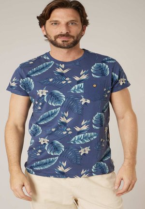 Homme barbu portant un t-shirt bleu à imprimé floral tropical et un pantalon beige clair, debout devant un fond neutre.