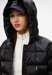 Czarna kurtka puchowa o pikowanym wzorze, z kapturem i sznurkami do regulacji. Noszona na białej bluzce z wysokim kołnierzem i zamkiem.