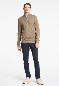 Beige strikket genser med halv glidelås, lange ermer og figursydd design, kombinert med mørke bukser og lyse sneakers.
