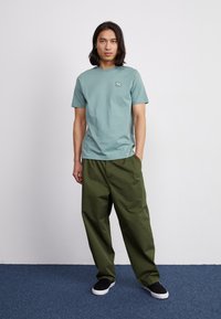 Tricou scurt cu mânecă scurtă de culoare teal deschis, cu un logo mic pe piept, asortat cu pantaloni largi de culoare verde măslin și pantofi negri slip-on.