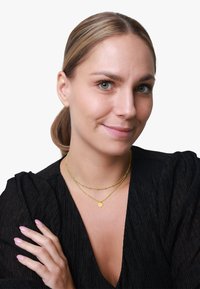 Zlatne ogrlice od slojeva sadrže okrugli privjesak i teksturirane perle, nošene preko crnog nabranog topa. Ten je svijetle boje. Ruke imaju bezbojni lak za nokte.