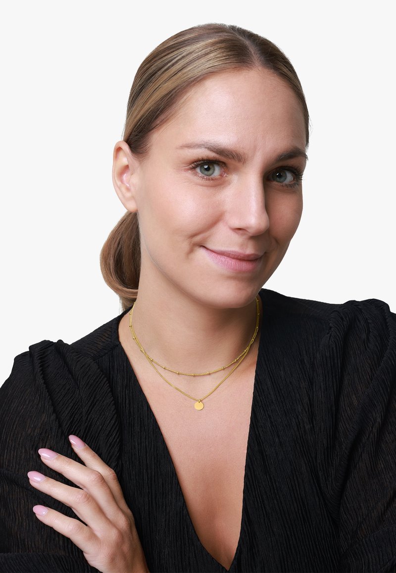 Zlatne ogrlice od slojeva sadrže okrugli privjesak i teksturirane perle, nošene preko crnog nabranog topa. Ten je svijetle boje. Ruke imaju bezbojni lak za nokte.