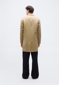 Άντρας που βλέπει προς τα πίσω, φοράει μπεζ παλτό μέχρι το γόνατο και φαρδιά μαύρα παντελόνια, σε απλό ανοιχτό φόντο.