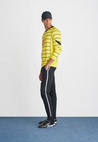 Κίτρινο μπλουζάκι μακρυμάνικο με μαύρες ρίγες και γραφικό σχέδιο, συνδυασμένο με μαύρη φόρμα joggers που έχει λευκή κάθετη ρίγα, και μαύρα παπούτσια.