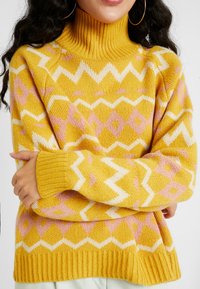 Żółty sweter z dzianiny z wysokim kołnierzem, ozdobiony białymi i różowymi wzorami zygzaków i diamentów. Mankiety i dolny brzeg z prążkowanej dzianiny dodają tekstury.