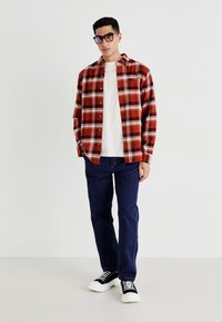 Camisa de cuadros roja y negra con botones blancos, sobre una camiseta blanca, combinada con jeans azul marino y zapatillas negras con suelas blancas.