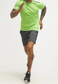 Conjunto de running que incluye una camiseta de manga corta texturizada en verde brillante y pantalones cortos oscuros con estampado. Zapatillas deportivas negras con detalles blancos.