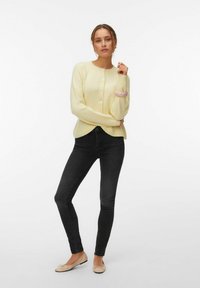 Femme debout les bras croisés, portant un cardigan boutonné de couleur jaune pâle, un jean skinny noir, des ballerines beiges et un bracelet rose.