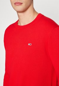 Roter Strickpullover mit rundem Halsauschnitt. Verfügt über ein kleines gesticktes Logo in Blau, Weiß und Rot auf der linken Brust. Klassische Passform und Textur.