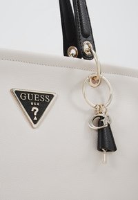Sac à main Guess en cuir blanc avec poignées noires, ferrures dorées, logo triangulaire et breloque en forme de clé suspendue à un anneau.